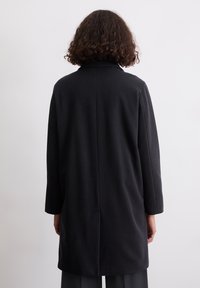 Person mit lockigem, dunklem Haar, der einen schwarzen langen Mantel trägt und vor einer schlichten weißen Wand steht.