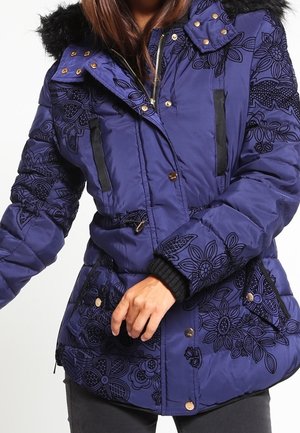 Kobieta w niebieskiej pikowanej kurtce z czarnymi kwiatowymi wzorami, czarnym kapturze z sztucznego futra i czarnych spodniach, poprawiająca przednią część kurtki.