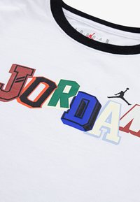 Camiseta de algodón blanca con letras "JORDAN" de colores en varios estilos de bloque; cuello acanalado negro; logotipo de Jordan en silueta negra.