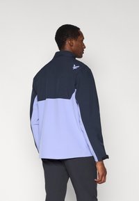 Jachetă pentru bărbați în culoarea bleumarin și mov deschis, cu guler înalt, mâneci lungi și manșete reglabile. Are un logo cu pasăre și un design în două nuanțe.