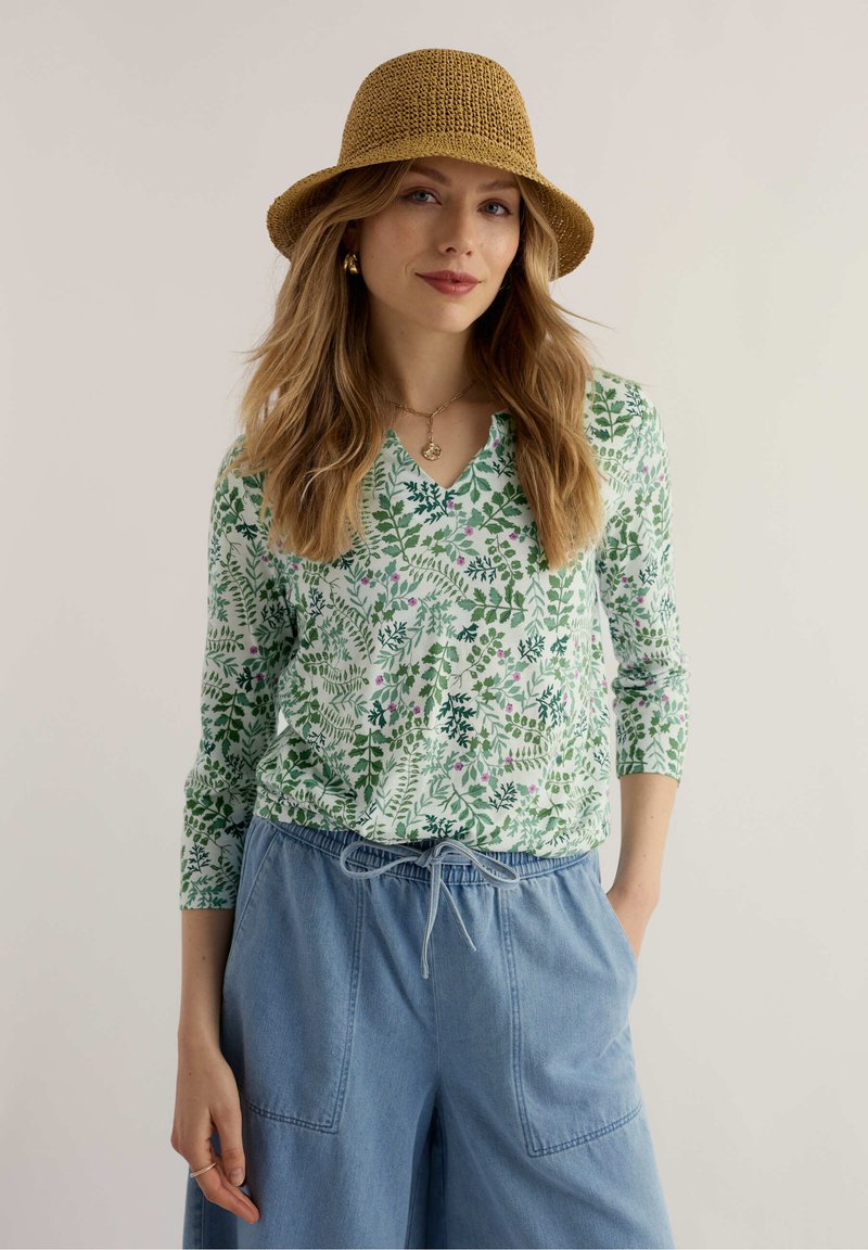 Frau mit einer senfgelben Strickmütze, grüner Bluse mit Blattmuster, blauen, hoch taillierten Hosen mit Kordelzug und goldenen Schmuckstücken, die ruhig steht.