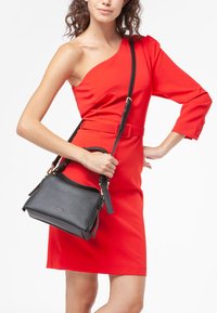 Borsa a mano in pelle nera con finitura textured, caratterizzata da una forma strutturata, manici doppi e una tracolla regolabile. Indossata con un vestito rosso.