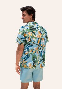 Koszula z krótkim rękawem, z niebieskim tłem i wyrazistymi zielonymi oraz kwiatowymi wzorami; zestawiona z jasnoniebieskimi szortami.