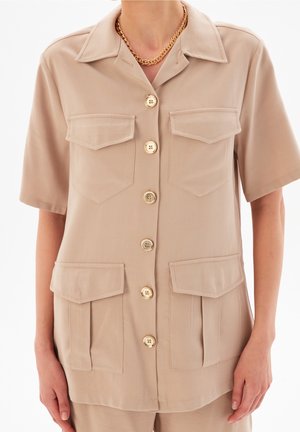 Beige overhemd met korte mouwen en knopen, voorzien van vier klepzakken, gedragen met een gouden ketting.