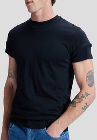 Mand iført en enkel sort t-shirt med opsmøgede ærmer og blå jeans med synlige tatoveringer på begge arme mod en neutral baggrund.