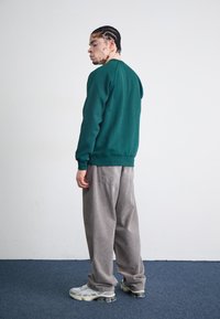 Green sweatshirt with a crew neck, gray corduroy trousers, and gray and beige sneakers. Smooth fabric and relaxed fit, standing against a plain wall.