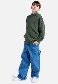 Grön huvtröja med framficka och snörning i huven, parat med löst sittande ljusblå denimjeans. Vita sneakers fulländar outfiten.