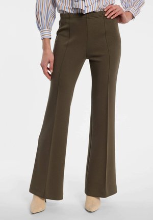 Person trägt hoch taillierte olivgrüne ausgestellte Hosen, gestreifte Bluse und beige spitze Schuhe, steht mit einer Hand in der Hüfte.