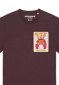 Burgunderfarbener Baumwoll-T-Shirt mit einer Grafik von Händen, die eine orangefarbene Kaffeetasse halten, umgeben von Wolken und Sternen. Der Text lautet "DER KAFFEE."