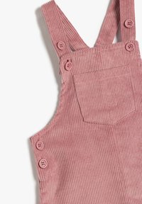 Pink corduroy overalls med justerbare stropper og to knapper på hver strop. Har en lille baglomme og lodrette ribbede strukturer.