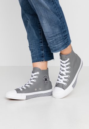 Personne portant des baskets montantes Converse grises avec des lacets blancs et des semelles à motif chevron, associées à un jean bleu raccourci.