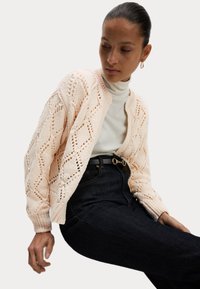 Hellrosa gestrickte Strickjacke mit Rautenmuster, gerippten Bündchen und offener Vorderseite, die über einem weißen Rollkragenpullover getragen und mit dunklen Jeans kombiniert wird.