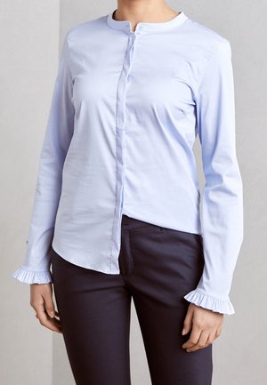 Kvinde iført en lyseblå bluse med lange ærmer og flæse manchetter samt marineblå tætsiddende bukser, der står foran en ensfarvet baggrund.