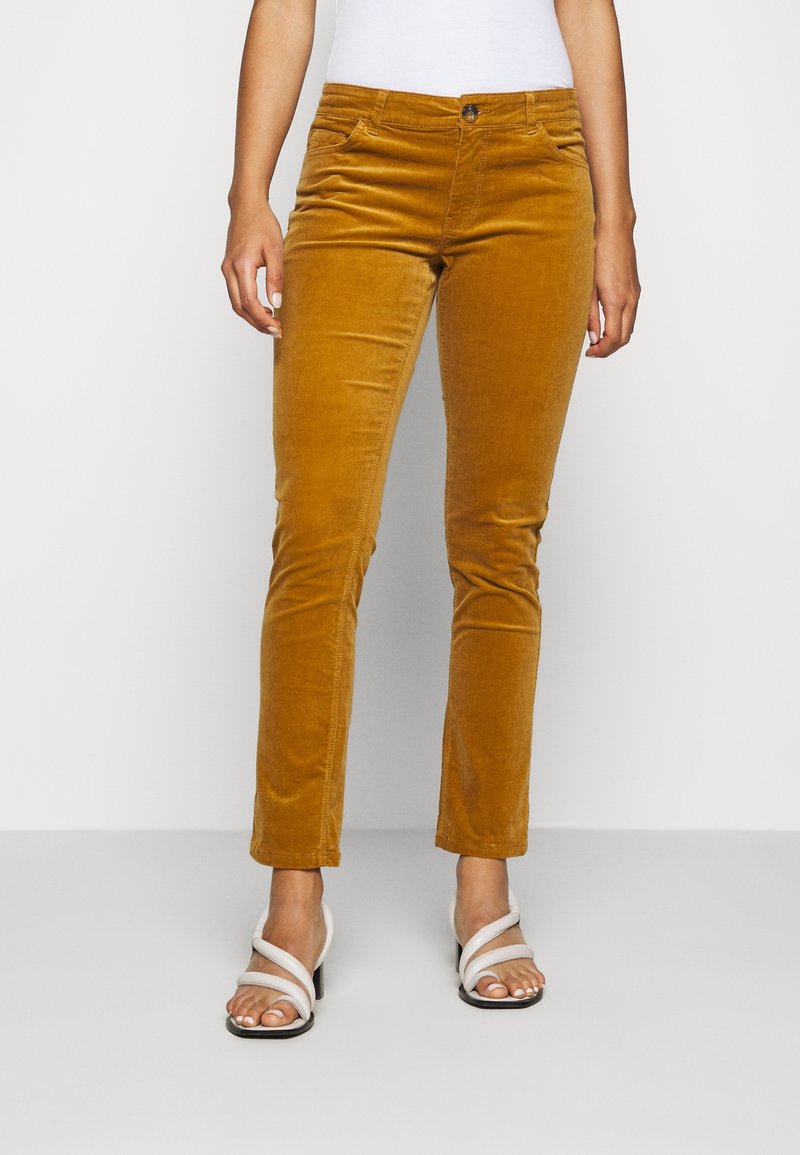 Femme portant un pantalon slim en velours côtelé jaune moutarde avec un bouton devant et des sandales blanches à talons avec des sangles croisées.