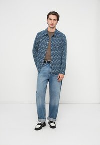 Geacă de blugi cu model zigzag albastru, combinată cu o cămașă maro și blugi bleu deschis. Pantofi negri și albi completează ținuta.