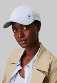 Lyseblå baseballcaps laget av stoff, med et lite sølv "ck"-logo, kombinert med en beige jakke og lagdelt lyseblå skjorte.