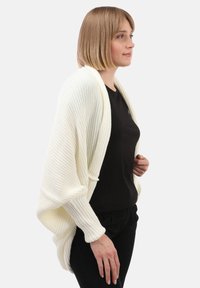 Frau mit kurzem blonden Haar, die einen cremefarbenen gestrickten Schal über einem schwarzen Oberteil und schwarzen Hosen trägt, mit der rechten Seite zu einem schlichten Hintergrund gewandt.