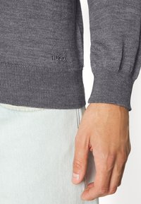 Grauer Strickpullover mit gerippten Bündchen und einem dezenten "HUGO"-Logo. Die Struktur wirkt glatt, getragen zu hellblauer Jeans.