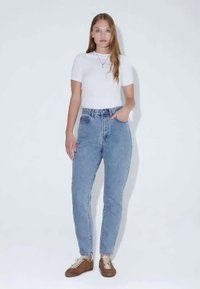 Ανοιχτό μπλε ψηλόμεσα τζιν με στενή γραμμή, μπροστινές τσέπες και θηλιές για ζώνη, συνδυασμένα με ένα εφαρμοστό λευκό μπλουζάκι και καφέ παπούτσια.