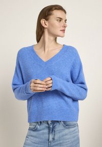 Helles Blau, weicher Strickpullover mit V-Ausschnitt, langen Ärmeln und geripptem Saum. Der Stoff wirkt strukturiert und gemütlich, in Kombination mit heller Jeans.