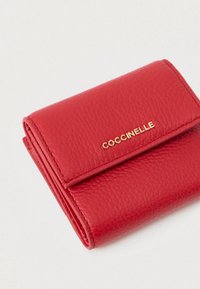Portafoglio in pelle rossa con texture, logo dorato "COCCINELLE" sul flap anteriore, piegato e posizionato su uno sfondo bianco.