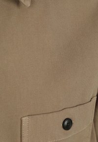 Beige stoffen zak met een zwarte knop, met een gestructureerde weving en ontwerp, die de functionele details van het kledingstuk benadrukt.