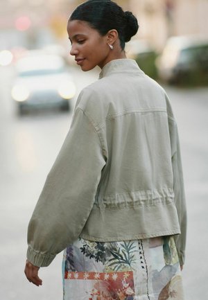 Young woman with dark hair in bun wearing hoop earrings, beige jacket, and floral patterned skirt, walking on an urban street at dusk.