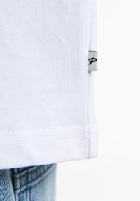 Hvid bomulds t-shirt med lige kant, der har et lille gråt mærke på venstre side, sammen med lyseblå denim jeans.