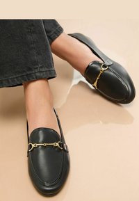 Schwarze Lederloafers mit einer abgerundeten Zehe, ausgestattet mit einem goldenen Kettenakzent und einem seitlichen Riemen, präsentiert vor einem neutralen Hintergrund.
