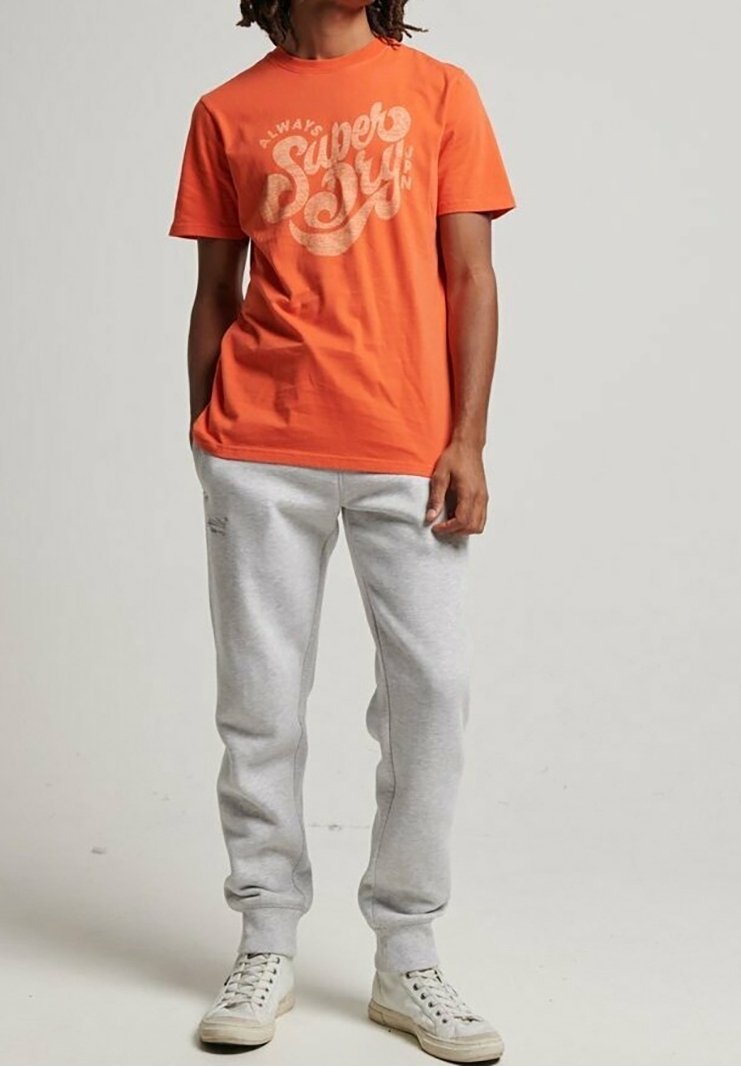 Orange bomulds t-shirt med hvid tekstureret "Superdry" grafik, parret med grå sweatpants og hvide sneakers. Letvægts, afslappet design.