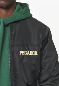 Giacca in nylon nera con chiusura a zip e logo ricamato "PEGADOR" in oro sul petto sinistro, indossata sopra un hoodie verde.