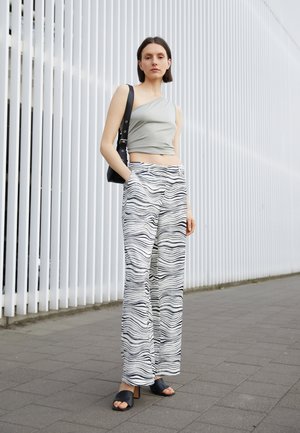 Top cropped cinza de um ombro, combinado com calças brancas de perna larga com um padrão ondulado preto. A modelo usa saltos abertos pretos e carrega uma bolsa preta.