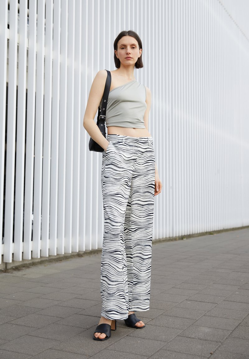 Top cropped cinza de um ombro, combinado com calças brancas de perna larga com um padrão ondulado preto. A modelo usa saltos abertos pretos e carrega uma bolsa preta.