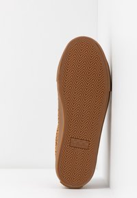 Suola di scarpa in gomma con un motivo a zigzag di colore marrone, caratterizzata da una superficie testurizzata e un'etichetta quadrata con logo vicino al tallone.