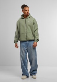 Green zip-up hoodie with front pockets, paired with loose-fitting light blue jeans and white sneakers, on a neutral gray background.
