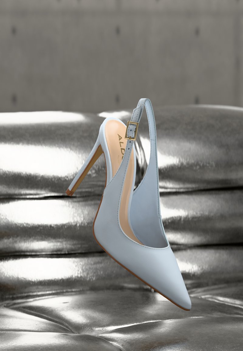 Scarpa slingback azzurro chiaro con tacco alto, punta appuntita e fibbia dorata, appoggiata su una superficie di pelle metallizzata argento lucido.