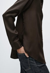 Camicia marrone in raso a maniche lunghe con davanti abbottonato e orlo curvo, abbinata a jeans in denim scuro. Texture liscia e vestibilità sartoriale.