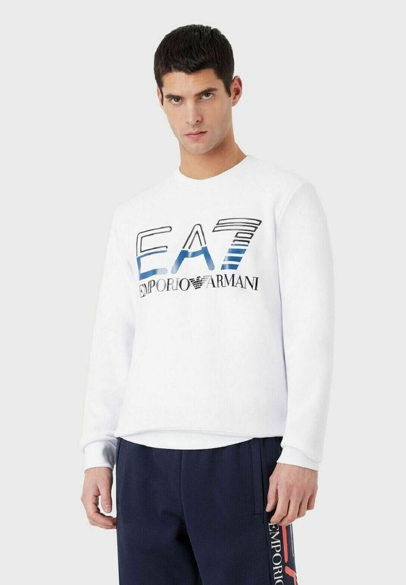 Felpa bianca con logo "EA7 Emporio Armani" blu e nero, scollo rotondo, maniche lunghe e polsini a costine. Abbinata a pantaloni blu scuro.