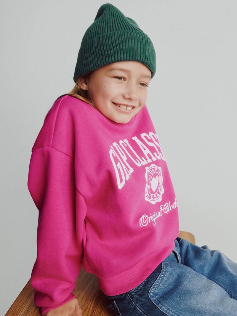 Felpa rosa brillante con logo bianco, abbinata a un berretto a coste verde. I jeans di denim casual completano l'outfit. Texture uniforme.