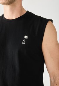 T-shirt en coton noir sans manches avec un design de palmier et de planche de surf brodé en blanc sur le côté gauche de la poitrine. Texture lisse.