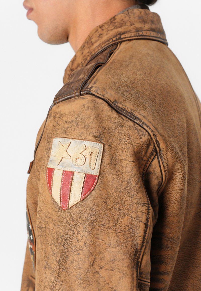 Giacca in pelle marrone invecchiata con finitura testurizzata, patte sulle spalle con design a scudo e accenti rossi e bianchi.