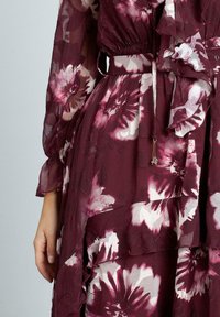 Vestito floreale color borgogna con tessuto trasparente a strati, caratterizzato da grandi motivi di fiori rosa e bianchi, vita arricciata e maniche lunghe e fluide.
