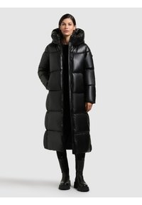 Giacca a vento nera con cappuccio, caratterizzata da una finitura lucida, design trapuntato e lunghezza lunga. Indossata sopra un outfit nero con stivali robusti.