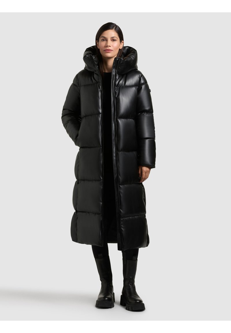 Giacca a vento nera con cappuccio, caratterizzata da una finitura lucida, design trapuntato e lunghezza lunga. Indossata sopra un outfit nero con stivali robusti.