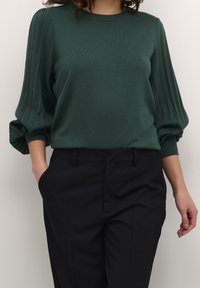 Pull en tricot vert avec des manches bouffantes à côtes, associé à un pantalon noir ajusté. Le tissu semble doux avec une texture lisse.