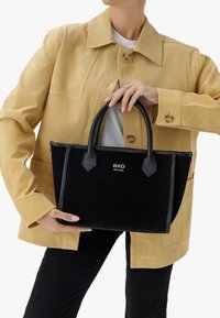 Czarna filcowa torebka z skórzanymi akcentami, podwójnymi uchwytami i wytłoczonym logo. Model nosi beżową zamszową kurtkę na białej bluzce i czarnych spodniach.