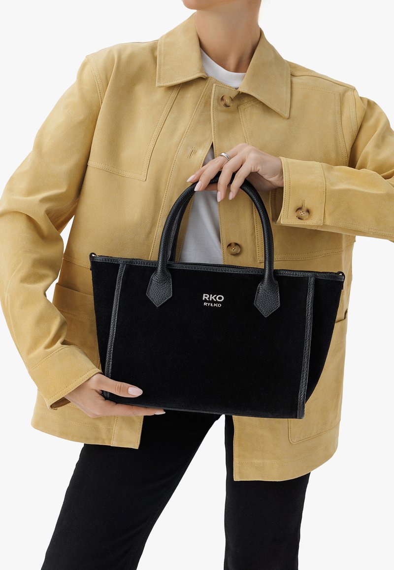 Czarna filcowa torebka z skórzanymi akcentami, podwójnymi uchwytami i wytłoczonym logo. Model nosi beżową zamszową kurtkę na białej bluzce i czarnych spodniach.