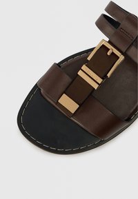 Καφέ δερμάτινο σανδάλι με μαύρη σόλα, που διαθέτει μεταλλικό κούμπωμα σε χρυσή απόχρωση στο κέντρο και σχέδιο με διπλή λωρίδα. Λείος υφιστάμενος και ανθεκτική κατασκευή.
