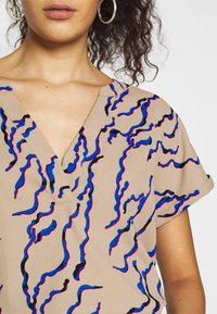 Béžový top s krátkým rukávem a výstřihem do V, zdobený plynulými modrými, červenými a černými vlnitými vzory na texturované látce.