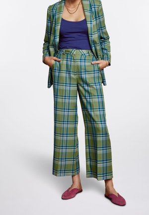 Femme portant un tailleur à carreaux verts et bleus avec un pantalon à jambes larges, un débardeur bleu marine, des colliers en or et des chaussures roses à enfiler, les mains dans les poches.